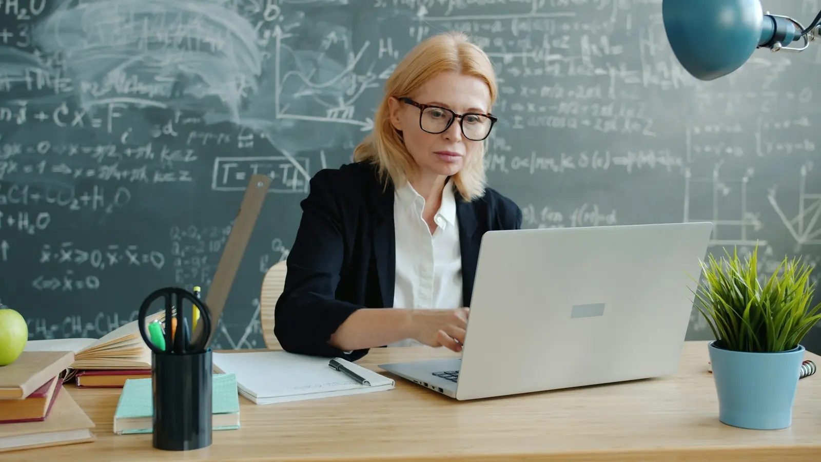 Professora trabalhando em um notebook diante de um quadro com fórmulas.