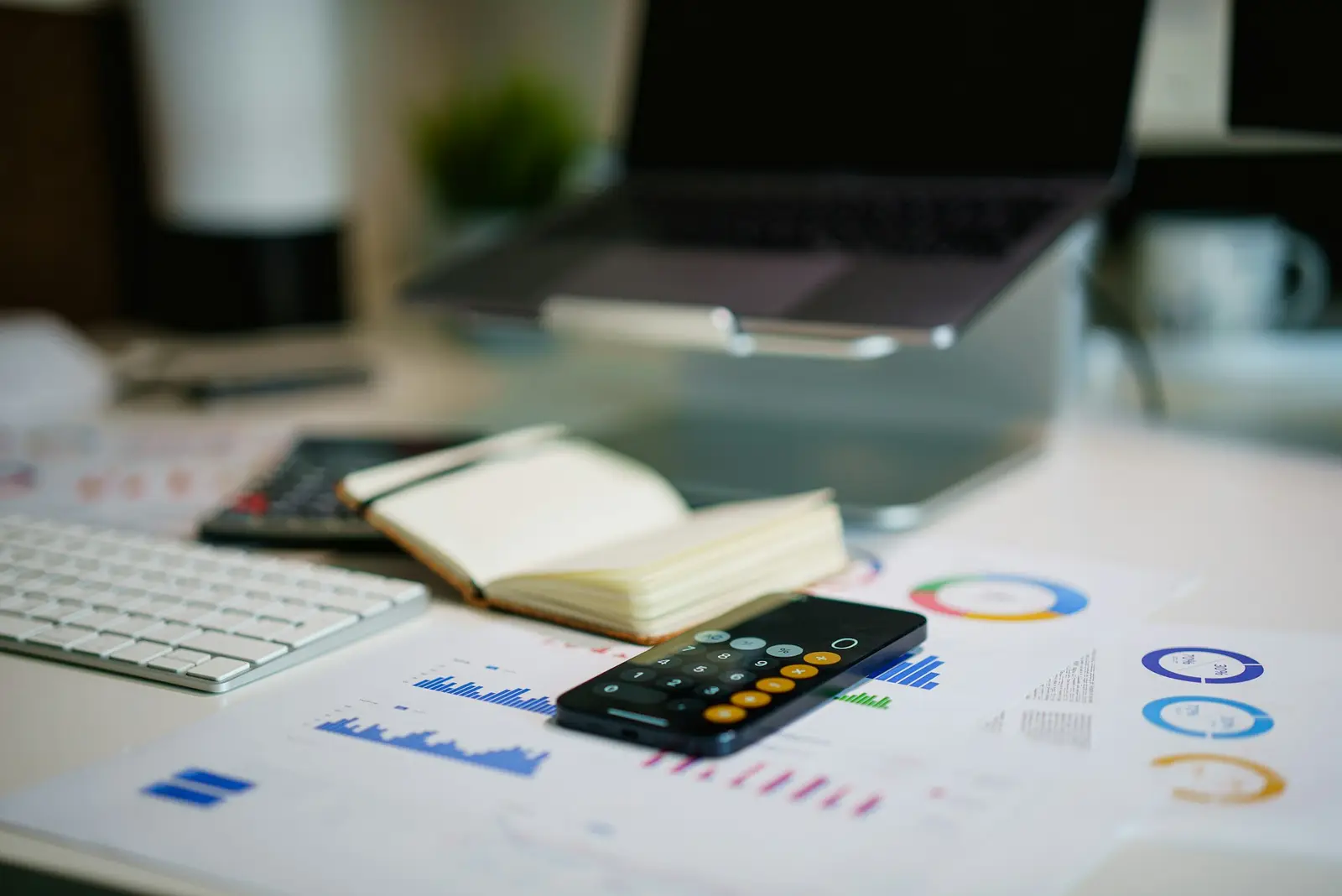 Mesa de escritório com calculadora no celular, caderno e gráficos financeiros impressos.