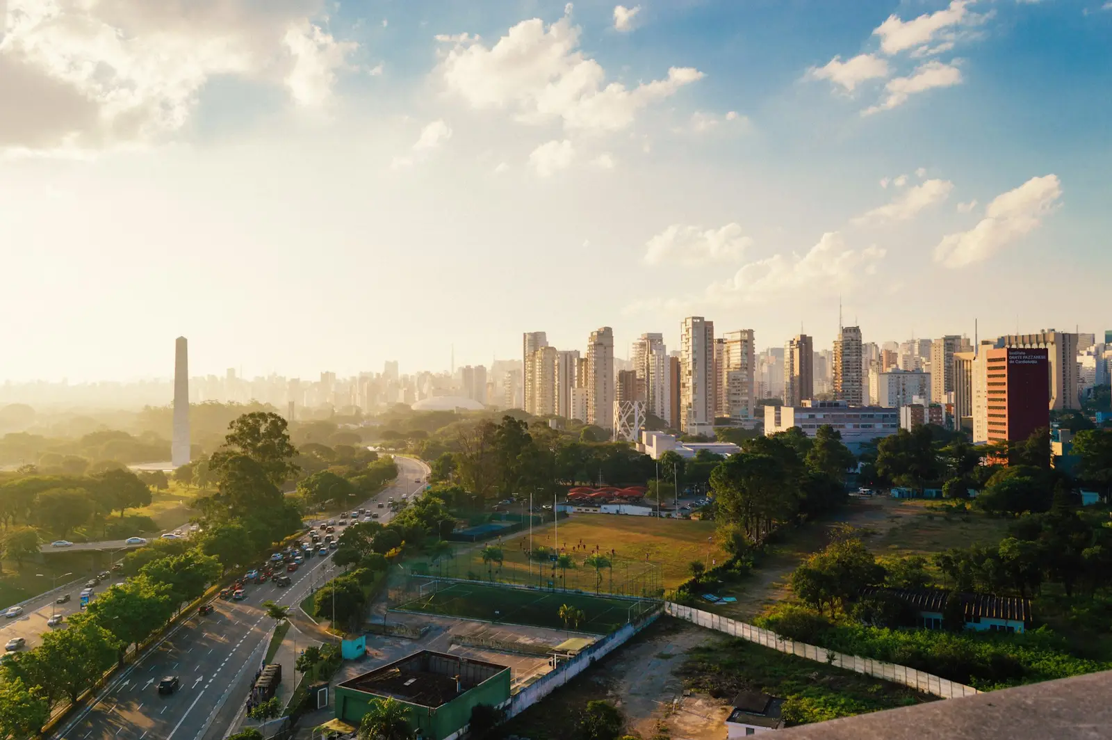 Vista do alto de uma cidade, com prédios, avenidas e um parque ao pôr do sol.
